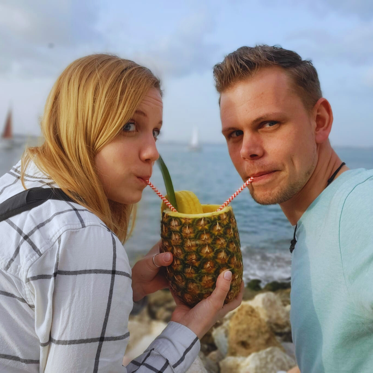 Olli und Julia von Hadscho Unterwegs trinken gemeinsam aus einer Ananas am Meer in Lissabon, Portugal, lachend in die Kamera
